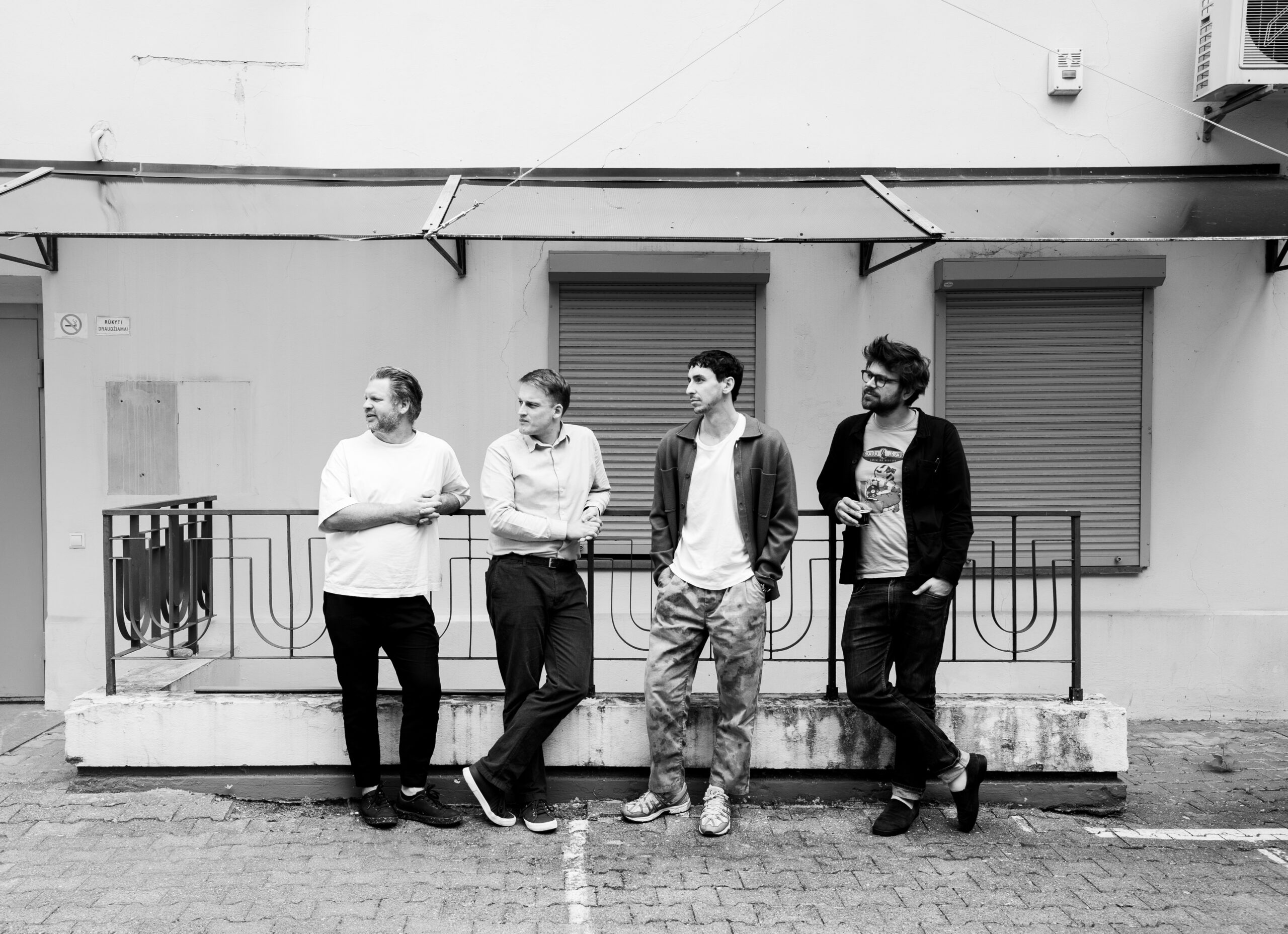 Four individuals standing casually in front of a minimalistic backdrop with modern railing details. The clean, structured environment reflects the contemporary design aesthetic found in exhibition spaces, retail stores, bars, hotels, and restaurants, emphasizing simplicity, functionality, and an urban architectural style. Parodų architektūra, parodų architektai, viešbučių, restoranų, barų, ir kavinių interjero dizainas